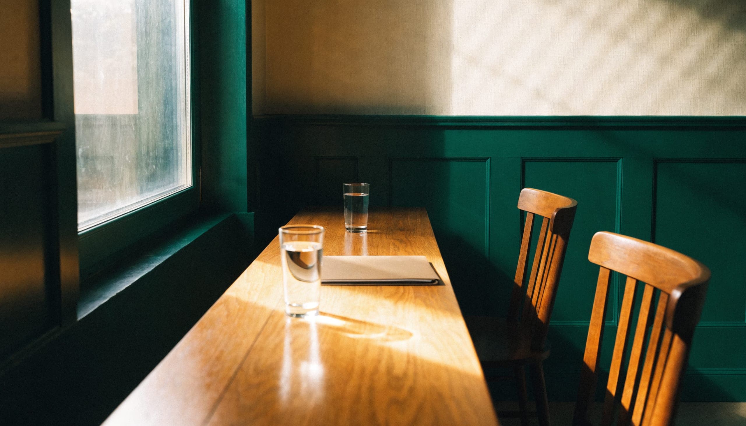 An empty meeting table by a tall window, two chairs pulled out and two glasses of water, in early morning light.